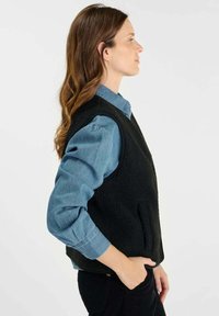 Gilet en polaire noire avec un col montant, des poches latérales et une coupe décontractée, porté par-dessus une chemise bleue à boutons, vu de côté.