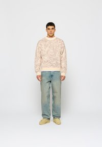 Svetlo ružový vzorovaný sveter s okrúhlym výstrihom, v kombinácii so svetlomodrými džínami. Modelka má na sebe béžové mokasíny.