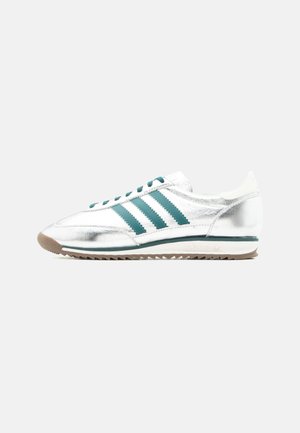 Hvid og sølv lavtop-sneaker med grønne striber, grønne snørebånd, hvid sål og gummiydersål, vist fra siden på en ensfarvet baggrund.