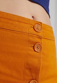 Oranžová džínová sukně s dvěma velkými, kulatými, vzorovanými knoflíky; viditelný prošívaný šev a strukturovaný detail látky kolem pasu.