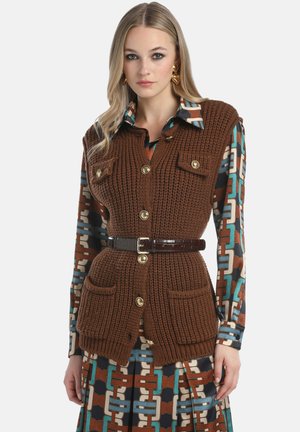 Brown knitted vest with gold buttons, two front pockets, and a textured finish, worn over a patterned shirt and complemented by a belt.