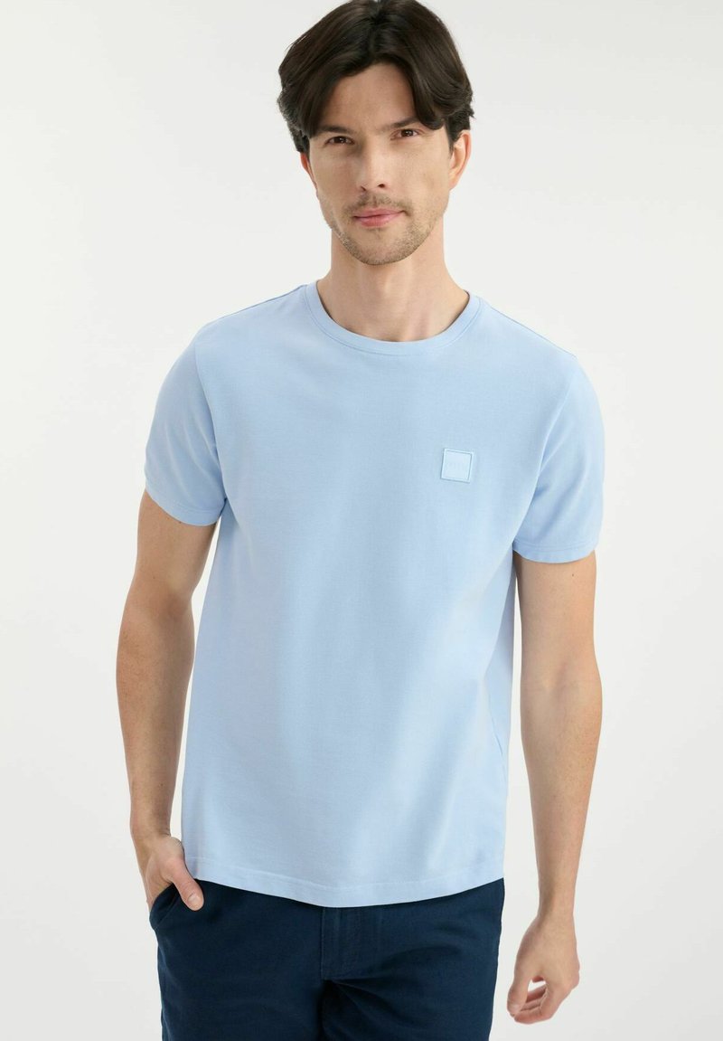 Jasnoniebieska koszulka z krótkim rękawem, wykonana z miękkiego materiału, z kwadratową haftowaną naszywką po lewej stronie klatki piersiowej oraz okrągłym dekoltem.