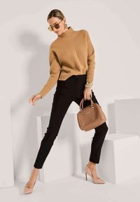 Frau in einem camel-farbenen, gerippten Pullover, schwarzen, schmalen Hosen, nudefarbenen Absätzen, goldenen Ohrringen und Armbändern, die eine braune geflochtene Handtasche hält und drinnen posiert.