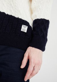 Paksu neulepaita vohvelinvärisessä ja laivastonsinisessä. Siinä on kaapeli-kuvioita, joustavat hihansuut ja pieni brändilogo laivastonsinisellä alueella.
