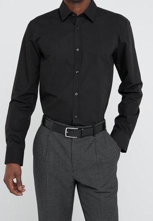 Mand iført en tætsiddende sort skjorte med knapper, stoppet ned i sort-hvide ternede bukser med sort bælte, hånd i lommen.