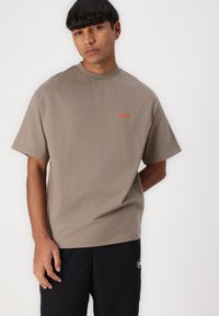 T-shirt in cotone marrone con collo rotondo e maniche corte. Presenta un logo arancione a contrasto "bareen" sul lato sinistro del petto. Texture liscia.