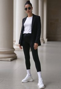 Černý blazer přes bílé tričko, spárovaný s černými legínami a bílými teniskami. Doplňky zahrnují kulaté sluneční brýle a náušnice ve tvaru kruhu.