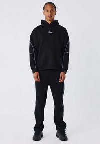 Joven de pie mirando al frente, vestido con una sudadera negra y pantalones negros a juego con finos adornos azul claro y zapatillas negras.