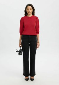 Top rosso aderente con maniche a sbuffo, abbinato a jeans neri a vita alta a gamba larga. Il modello tiene una piccola borsa nera e indossa tacchi neri.