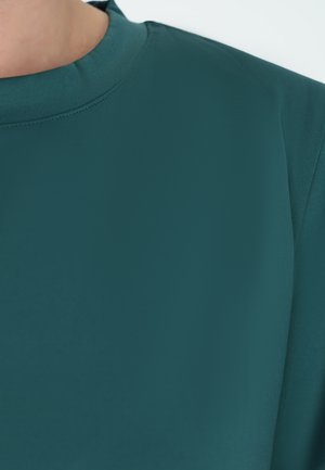 Teal blouse met een ronde halslijn, een gladde textuur en zachte stof. Close-up benadrukt de stiksels langs de kraag en schoudernaden.
