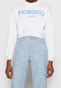Sweat-shirt blanc court avec texte "Fiorucci Milan" en bleu clair, associé à un jean en denim bleu clair orné d'un motif de logo répété.