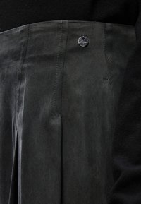 Calças plissadas verde-escuro com textura suave, apresentando um botão de logótipo prateado e detalhes de costura na cintura e nas costuras laterais.