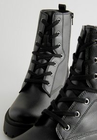 Zwarte enkellaarzen van gestructureerd leer met een vetersluiting, metalen oogjes en een chunky rubberen zool met een subtiele hak.