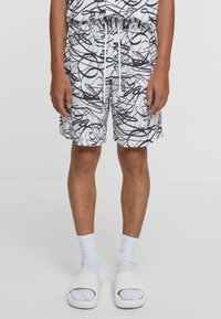 Short blanc avec motif abstrait tourbillonnant noir, taille élastique, cordon de serrage et poches latérales. Associé à des sandales blanches à enfiler et des chaussettes.