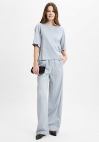 Vrouw staat met een losse lichtblauwe top en brede broek, houdt een zwarte clutch vast, draagt zwarte spitse schoenen en een dunne ketting.