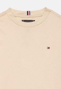 Béžové tričko vyrobené z bavlny, s okrúhlym výstrihom a malým vyšitým logom v modrej, červenej a bielej farbe na hrudi.