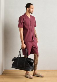 Burgundiškai tekstūruotas trumpomis rankovėmis marškiniai ir šortai derinami su smėlio spalvos šlepetėmis; laiko juodą sportinį krepšį su odiniais akcentais.