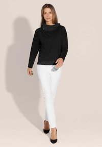 Zwarte longsleeve top met hoge kraag, voorzien van knopenaccenten, gecombineerd met slim-fit witte broek en zwarte hakken, tegen een neutrale achtergrond.