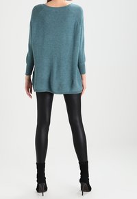 Pull en tricot surdimensionné turquoise avec des poignets côtelés, associé à des leggings noirs brillants et des bottines à talons noires. Texture lisse, coupe décontractée.