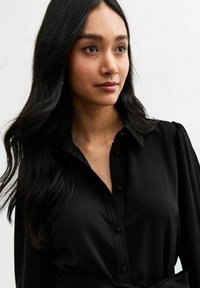 Camicia nera con bottoni, colletto, maniche lunghe e una texture liscia. Presenta una vita con cintura e dettagli sottili sulle spalle.