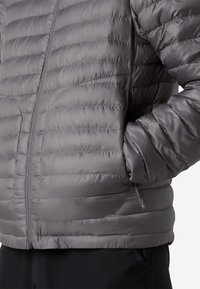 Grå pufferjacka med horisontellt quiltat mönster, zippered front, sidofickor och slät textur. Lätt och isolerad för komfort.