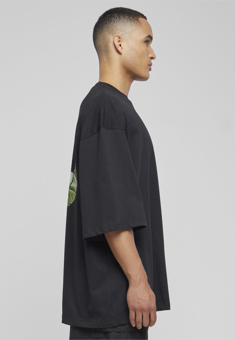 Giovane uomo con capelli corti in piedi di profilo, indossa una maglietta nera larga e oversize con una grafica verde sulla schiena.