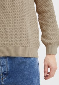 Beige neulepaita, jossa on teksturoitu timanttikuosi, resorit hihoissa ja helmassa. Yhdistetty sinisiin farkkuihin. Huomaamaton logo näkyy sivussa.