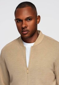 Beżowa kurtka bomberka o teksturowanym wykończeniu, z zapięciem na zamek i okrągłym kołnierzykiem, noszona na białej koszuli, na neutralnym tle.