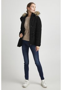 Zwarte, geïsoleerde jas met een faux fur-voering in de capuchon, een ritssluiting aan de voorkant en verstelbare koordjes, gecombineerd met een beige kabelgebreide trui en blauwe jeans.