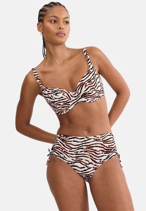 Frau mit geflochtenem Haar, die einen hoch taillierten Bikini mit schwarz-orangem und weißem Tigerstreifenmuster trägt, steht mit den Händen hinter dem Rücken.