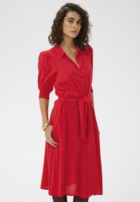 Robe rouge boutonnée avec manches courtes bouffantes et ceinture à nouer, dotée d'une jupe fluide et de poches latérales. Tissu lisse et léger.