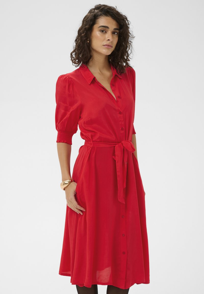 Robe rouge boutonnée avec manches courtes bouffantes et ceinture à nouer, dotée d'une jupe fluide et de poches latérales. Tissu lisse et léger.
