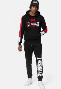 Sudadera negra con capucha y acentos en rojo y blanco, que presenta un logo; combinada con pantalones de chándal negros que exhiben un logo blanco en la pierna.