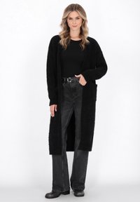 Cardigan lung negru, dintr-un material moale și pufoș, cu deschidere frontală, umeri lăsați și două buzunare frontale; purtat peste un top negru și blugi închiși la culoare.