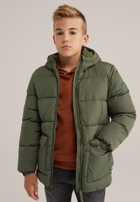 Zeleni puhovka s kapuco, zadrgo in okrasno nalepko, nosena čez rjavo pot sweatshirt. Ima elastične manšete in žepke.