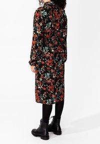 Vestito midi floreale con sfondo nero, caratterizzato da fiori arancioni, verdi e bianchi, maniche lunghe e vita cintrata. Indossato con stivali neri.