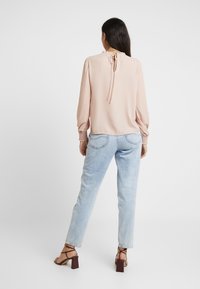 Rosa blus med rynkad halsringning och knytning i ryggen, ihop med ljusa blå jeans och bruna blockklackade sandaler. Slät tygkvalitet och åtsittande ärmar.