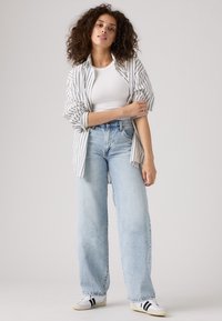 Model fehér bordázott trikóban, túlságosan nagy csíkos ingben és világoskék, széles szárú, laza szabású farmerral, fekete-fehér tornacipővel párosítva.