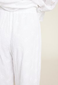 Hvite broderte bukser laget av lett stoff. Har øyelette mønstre og elastisk midje. Glatt tekstur med løs passform.