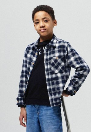 Marineblå og hvid ternet flannel skjorte med knapper, parret med en sort sweatshirt og blå jeans. Bemærkelsesværdig lomme detalje på skjorten.