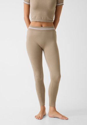 Persoon die beige aangesloten leggings en bijpassende cropped top met korte mouwen draagt, blootsvoets staand tegen een effen lichte achtergrond.
