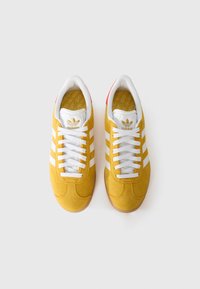 Gele suede sneakers met drie witte strepen, witte veters en een gumrubberen zool. Het interieur heeft een geel patroon in de inlegzool.