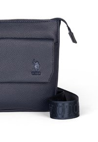 Navy modrá texturovaná kožená taška se zipovým uzávěrem, přední kapsou a popruhem s embosovaným logem. Kompaktní a funkční design.
