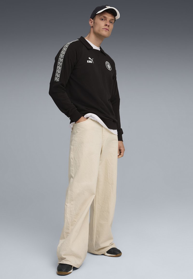 Polo negru cu mânecă lungă, având logo alb și accente cu model pe mâneci, asortat cu pantaloni crem, evazați, și pantofi negri.