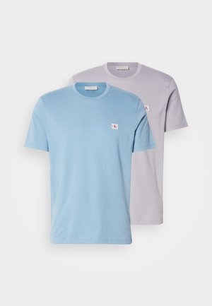 Deux t-shirts col rond : un bleu clair, un gris clair. Tous deux sont à manches courtes, fabriqués en tissu doux, et possèdent un petit logo sur la poitrine.
