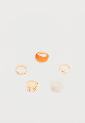 Cinq bagues assorties posées sur une surface blanche, présentant des designs en or, orange translucide et marbré blanc.