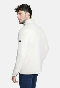 Chaqueta de plumas blanca con cuello alto, que presenta un detalle de botón en la manga y una textura suave. Lleva sobre pantalones azul oscuro.
