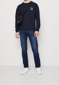 Sudadera azul marino con cuello redondo y parche gráfico, combinada con jeans de mezclilla azul y zapatillas blancas. Bolsa bandolera de material negro.