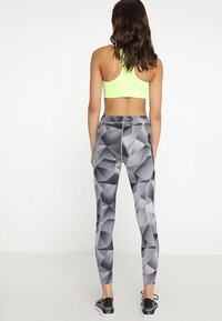 Nainen seisoo selin, päällään neonkeltainen urheiluliivi, harmaat geometriset leggingsit ja mustat urheilukengät.
