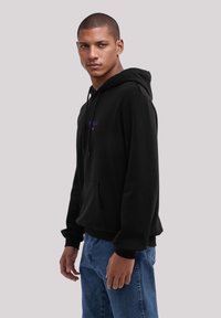 Sudadera negra con bolsillo frontal, capucha con cordones y texto morado. El modelo lleva puestos unos vaqueros azules, mostrando un ajuste relajado y un estilo casual.